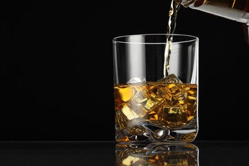 Pouring whiskey into glass with ice cubes at table against black background, space for text