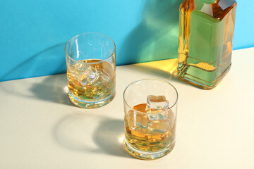 Whiskey with ice cubes in glasses and bottle on white table against light blue background