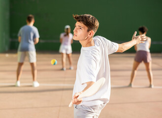 Portrait of sporty young guy playing pelota on open fronton court on summer day, ready to hit...