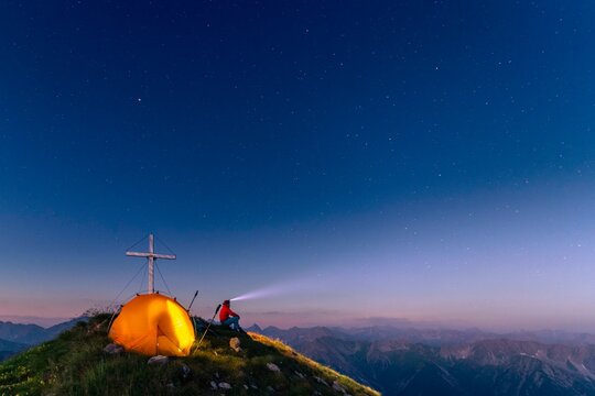 Cross of the Kreuzspitze by a starry sky with tent and mountaineer at the summit, in the background the Lechtal Alps, Elmen, Lechtal Alps, Ausserfern, Tyrol, Austria, Europe