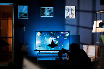 Player participates in online roleplaying firing match, hooking console through TV and enjoying nighttime gaming activity with friends. Male student is playing shooter video games. - obrazy, fototapety, plakaty