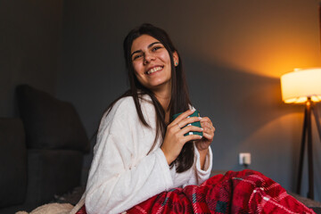 A young girl or woman getting up from bed drinking coffee or tea in blanket cozy room	