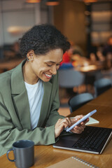 A woman sits at a table with a laptop and tablet, smiling in her blazer
