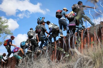 Border crossing: group of migrants climbing over fence with barbed wire.