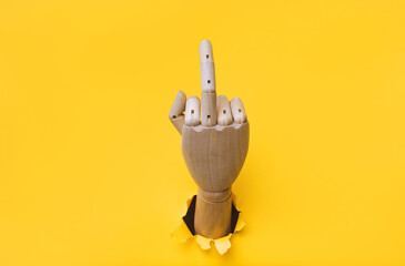 Middle finger of a right wooden hand, offensive gesture. Torn hole in yellow paper. Fuck you concept. Aggressive reaction of artificial intelligence or robot.