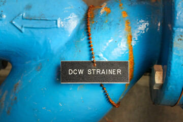 DCW Strainer label on pipe in water meter room. Close up of blue pipe with name tag and arrow to the domestic cold water strainer. Used to filter and remove particles from water. Selective focus.