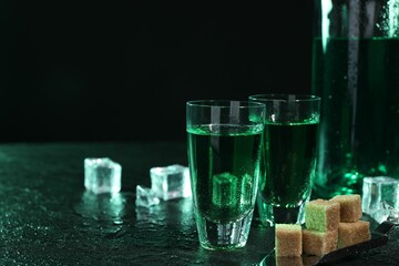 Absinthe in shot glasses, spoon, brown sugar and ice cubes on gray table against dark background,...