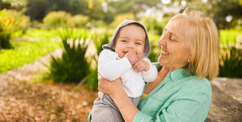 Fototapeta premium Beautiful happy smiling senior elderly woman holding on hands cute little baby boy. Grandmother and grandson having fun time together at tropical summer day in park. Multigenerational family concept