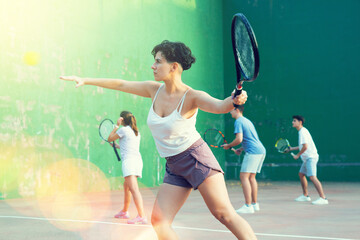 Sporty young Argentinian woman playing popular team match of frontenis at open-air fronton on summer day, ready to hit rubber ball with racquet