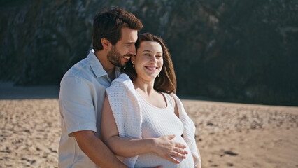 Smiling man embracing pregnant belly wife standing sandy seashore close up.