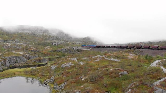 Aerial: ore train in S&oslash;sterbekk stasjon, close to the border between Sweden and Norway in north Lapland crossing a bridge on a lake and entering a tunnel in a misty landscape
