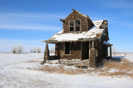 Outdoor winter rural prairie landscape scene of the exterior of an old, abandoned and rundown wooden farmhouse and veranda with a blue sky and snow