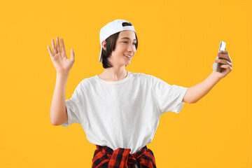 Cool teenage boy with mobile phone waving hand on yellow background