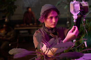 Medium shot of young Caucasian woman living in post-apocalyptic shelter watering plants with use of handmade filter system