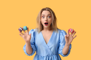 Shocked young woman with different sweet macaroons on yellow background