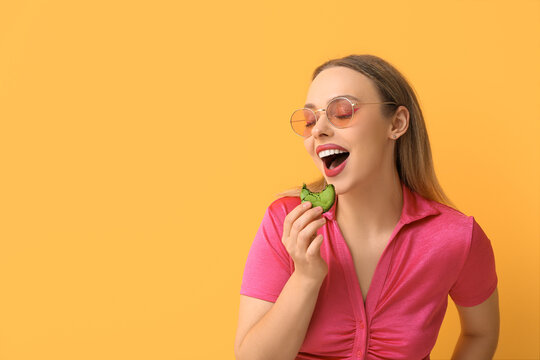 Beautiful young woman eating green sweet macaroon on yellow background