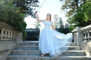 Fototapeta premium full length portrait of beautiful female model wearing blue fantasy ballgown, like a fairytale elf princess. Holding. Sword weapon, standing on staircase of a romantic castle balcony location.