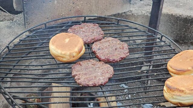 Close up of deer burgers being grilled alongside buns over fire pit burning wood sticks.