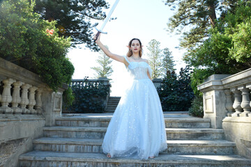 Obraz premium full length portrait of beautiful female model wearing blue fantasy ballgown, like a fairytale elf princess. Holding. Sword weapon, standing on staircase of a romantic castle balcony location.