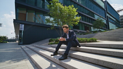 Successful businessman scrolling mobile phone sitting stairs urban street.