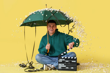 Young man with umbrella, movie clapper and film sitting on yellow background