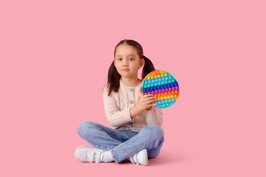 Little Asian girl with autistic disorder and pop it fidget toy sitting on pink background