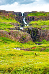 Fototapeta na wymiar Cover page with cascading waterfall in East Iceland, with dramatic sky and green meadow hills. Beautiful Icelandic landscape, huge cliff, water cascade and rock in moss
