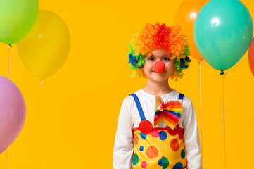 Funny little boy in clown costume with balloons on yellow background. April Fools Day celebration