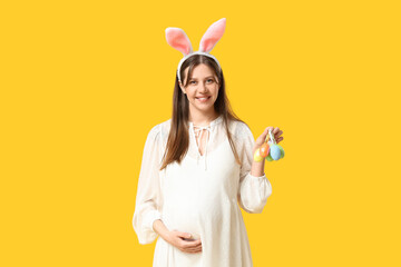 Beautiful young pregnant woman in bunny ears with Easter eggs on yellow background