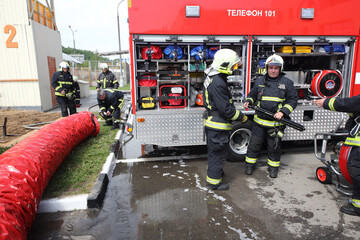 Five fireman working with large tube near fire truck with equipment on territory of fire station.