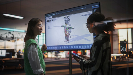 Black Female Game Designer Talking to Asian Female Project Manager And Standing Next To TV Set With...