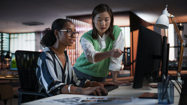 Asian Female Creative Director Giving Instructions on 3D Visuals to Black Female VFX Animator in Creative Agency Modern Office. Two Stylish Diverse Women Discussing Design on Desktop Computer