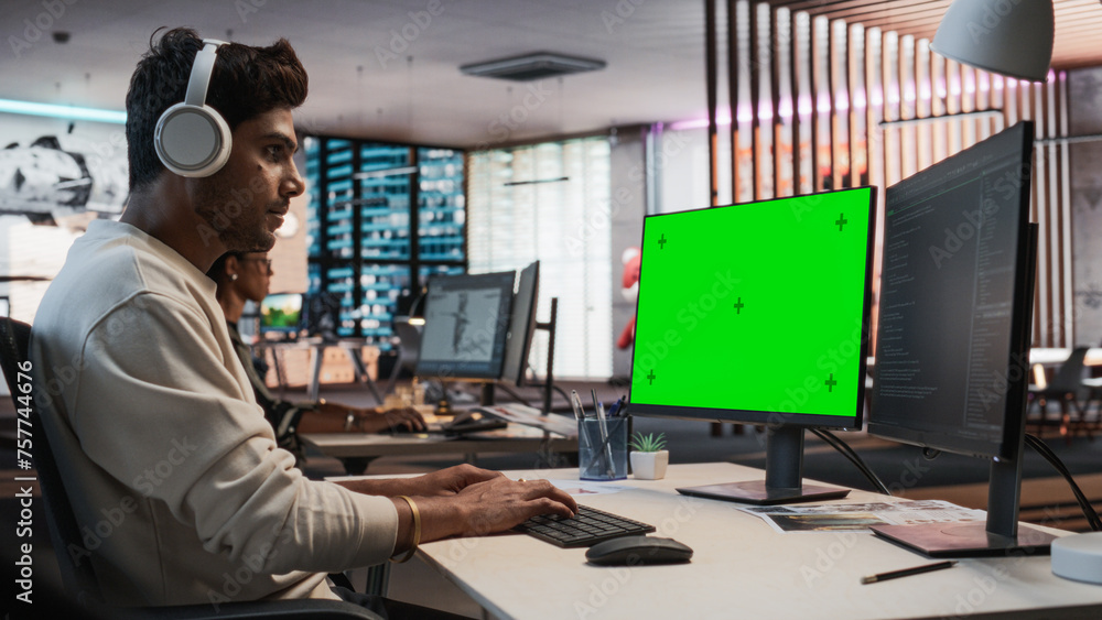 Poster Portrait of Creative Indian Man Sitting at a Desk Using Desktop Computer with Mock-up Green Screen Chromakey. Male Level Designer Working in Game Design Startup Office, Creating Immersive Gameplay.