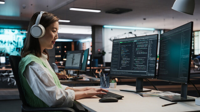 Female Asian Game Programmer Coding On Desktop Computer In Game Development Studio Diverse Office. Focused Woman Writes Lines Of Code, Does Gameplay Engineering For New Immersive 3D RPG Video Game.