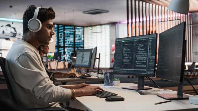Male Indian Game Programmer Coding On Desktop Computer In Game Design Studio Diverse Office. Focused Man Writes Lines Of Code, Does Gameplay Engineering For New Immersive 3D Survival Video Game.