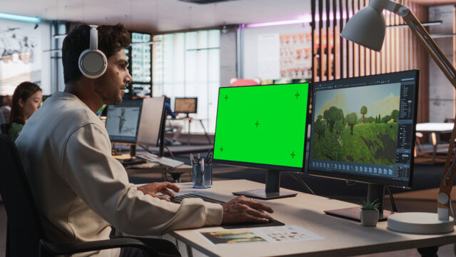 Indian Male Game Designer Using Desktop Computer With Green Screen Chromakey on Display, Designing Immersive World In 3D modelling Software For RPG Video Game. Man Working In Game Development Office
