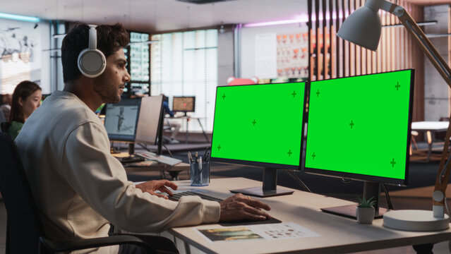 Indian Male Game Designer Using Desktop Computer With Green Screen Chromakey on Display, Designing Immersive World In 3D modelling Software For RPG Video Game. Man Working In Game Development Office