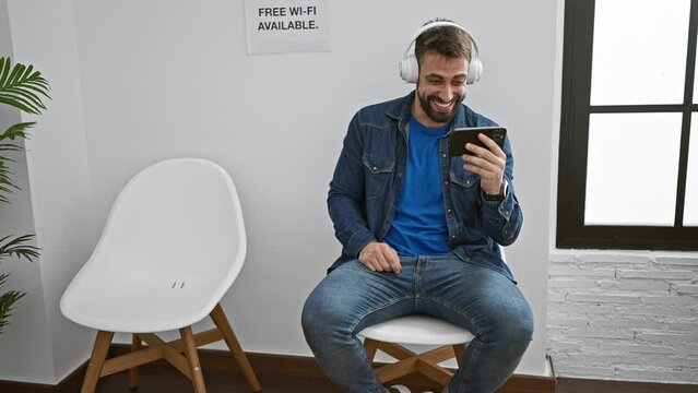 Handsome young hispanic man confidently immersed in vivid audio-visual world, sitting in waiting room chair, chilling, smiling as he's watching video and listening to music on smartphone.