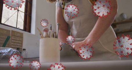 Image of covid 19 cells over woman working in pottery studio wearing face mask