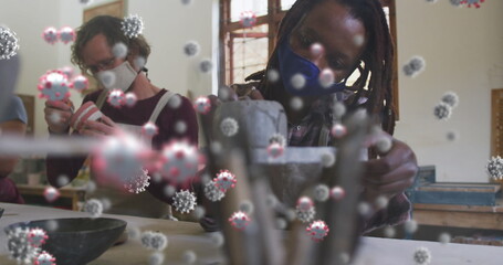 Image of virus cells over diverse workers with face masks painting pottery