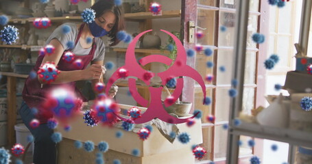 Image of virus cells and biohazard symbol over caucasian woman with face mask forming pottery