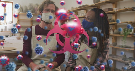 Image of virus cells and biohazard symbol over diverse workers with face masks