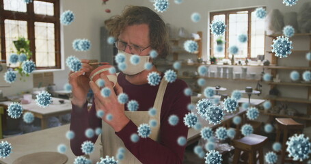 Image of covid 19 cells over man working in pottery studio wearing face mask