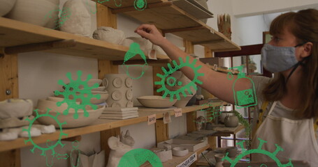 Image of virus icons over caucasian woman with face mask inspecting pottery