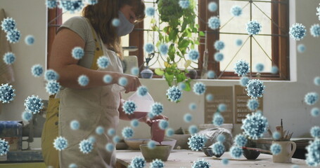 Image of covid 19 cells over woman working in pottery studio wearing face mask