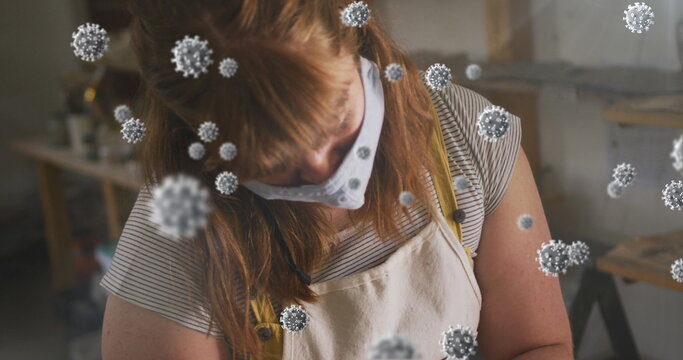 Image of covid 19 cells over woman working in pottery studio wearing face mask