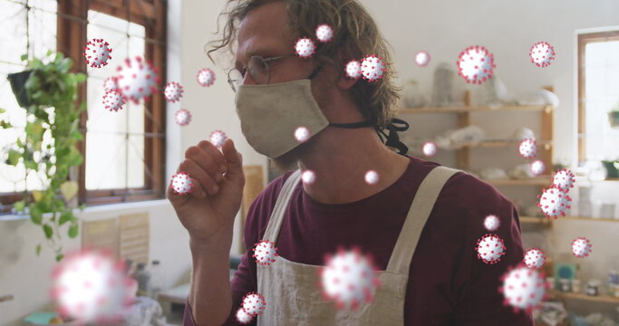 Image of coronavirus cells floating over caucasian man in face mask coughing at pottery workshop - Powered by Adobe