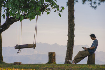 landscape and travel concept with solo freelancer man use tablet work at wooden swing under large...