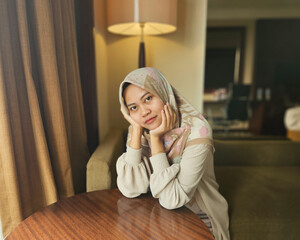 Asian Muslim woman wearing hijab sitting pensively while supporting chin and head, looking at...
