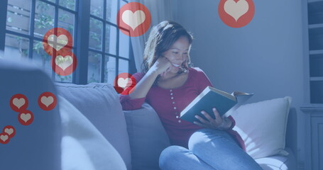 Image of digital heart icons over woman reading book and smiling at home